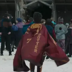 Chłopiec w pelerynie z napisem "POTTER 7" idzie przez śnieg w otoczeniu innych uczniów, jak w serialu Harry Potter.