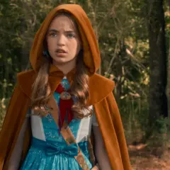 Holly Wheeler z Stranger Things w brązowym płaszczu z kapturem i niebieskiej sukience, stoi w lesie.