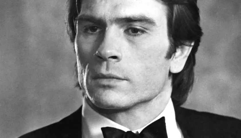 Młody Tommy Lee Jones w czarnym smokingu z muszką, zamyślony.