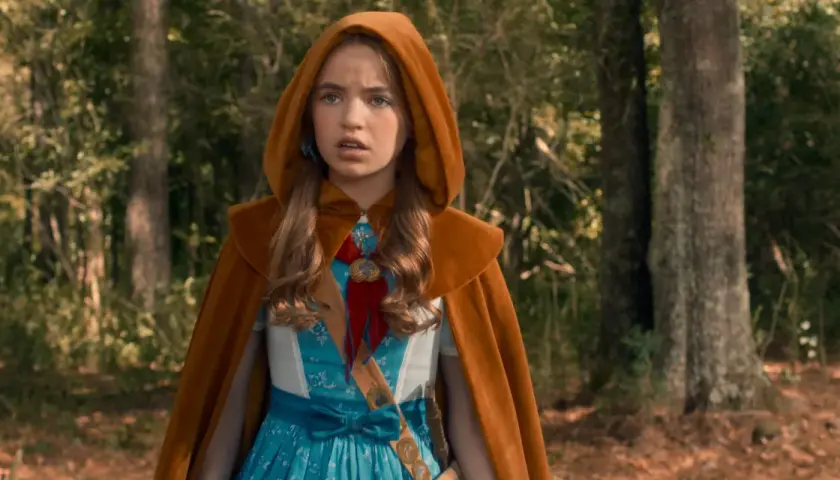 Holly Wheeler z Stranger Things w brązowym płaszczu z kapturem i niebieskiej sukience, stoi w lesie.