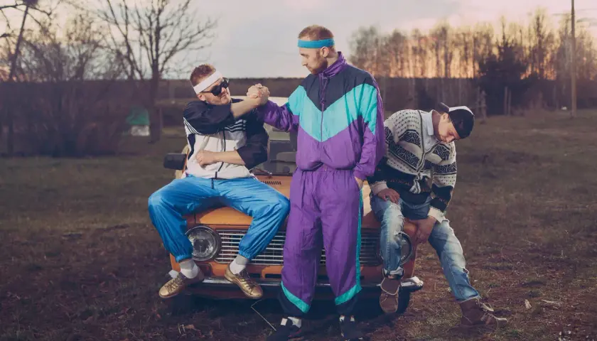 Trzech mężczyzn w strojach z lat 90. moda, siedzących na starym samochodzie. Jeden w dresie, drugi w bluzie i jeansach.