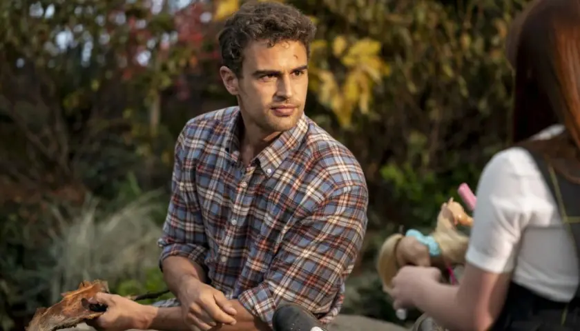 Theo James w kratowanej koszuli, znany z filmów, seriali i programów, siedzi na zewnątrz. Obok niego dziecko bawi się lalką.