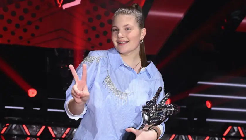 Młoda artystka z pucharem i znakiem pokoju, radośnie świętuje zwycięstwo w The Voice Kids.