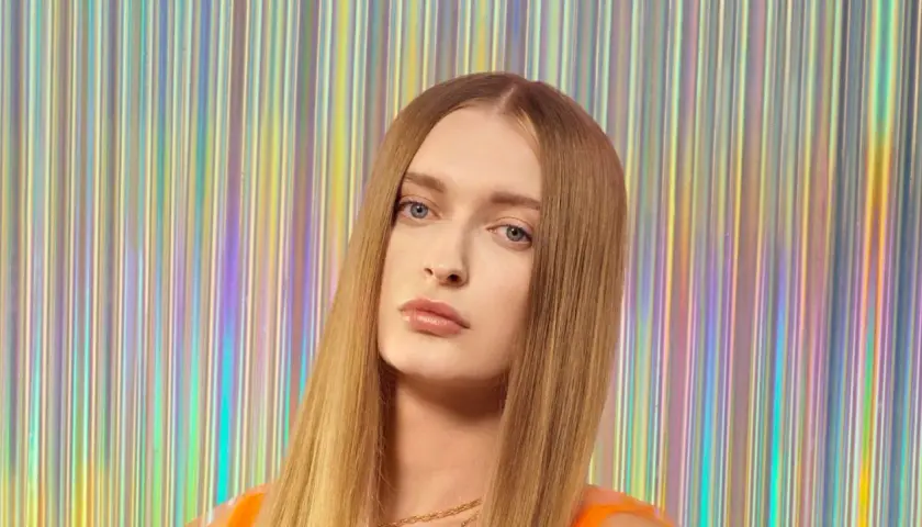 Sophie, top model o długich, blond włosach, pozuje na tle błyszczącej, tęczowej kurtyny.