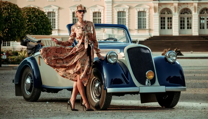 Elegancka kobieta w kwiecistej sukience i okularach przeciwsłonecznych opiera się o zabytkowy kabriolet. Styl lat 20. moda damska.