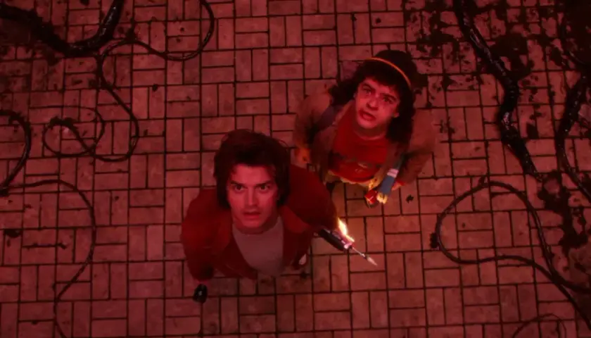 Steve i Dustin patrzą w górę, gotowi na ostateczną walkę w Stranger Things final. Ciemne macki wiją się na czerwonej posadzce.