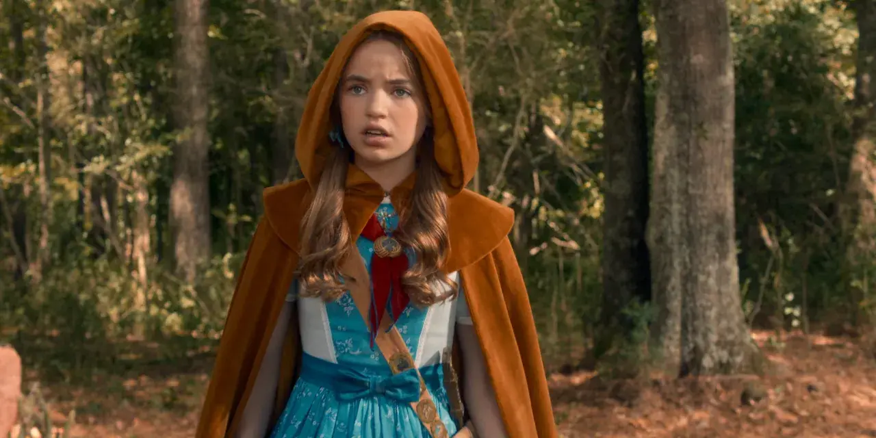Holly Wheeler z Stranger Things w brązowym płaszczu z kapturem i niebieskiej sukience, stoi w lesie.