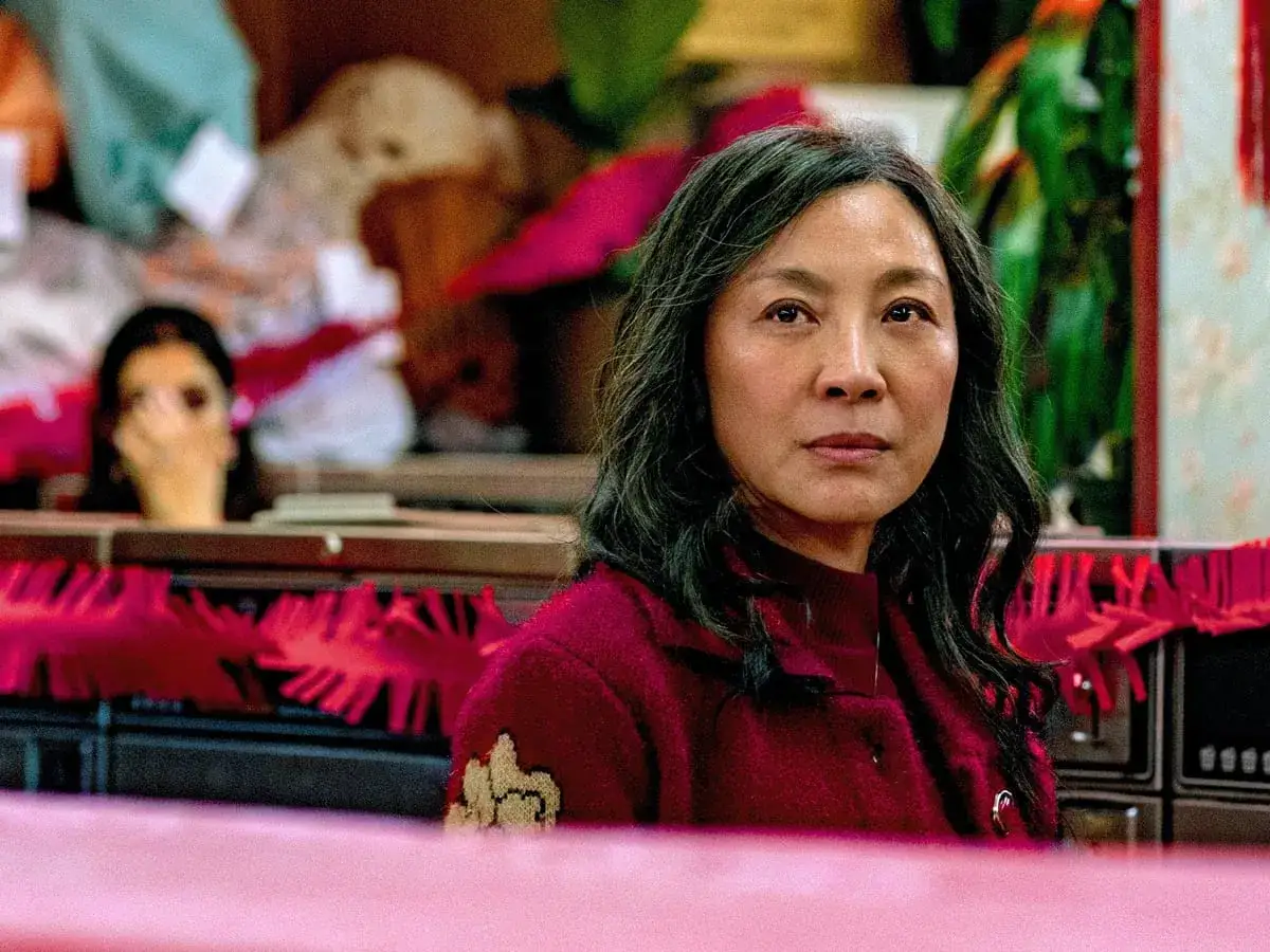 Kobieta w czerwonym swetrze, otoczona chaosem, jakby wszystko wszędzie naraz. W tle widać inną postać.